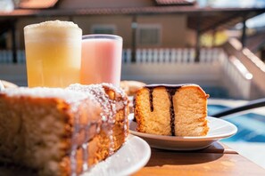 Una rebanada de pastel con chocolate derramado encima, un vaso de jugo de naranja y un vaso de bebida rosa sobre una mesa, con una piscina al fondo.