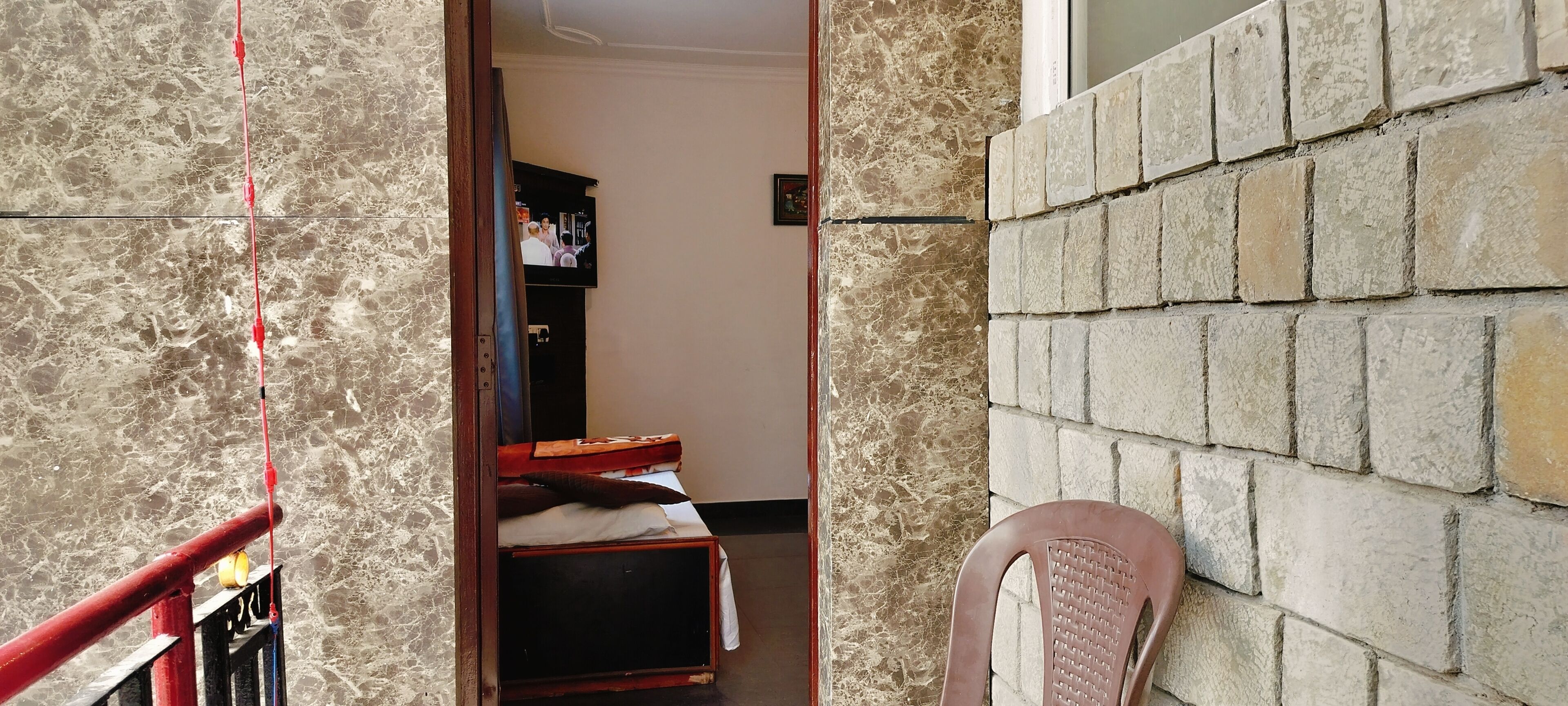 Un couloir avec un mur de marbre, une rambarde rouge, un lit recouvert d’un drap blanc, une chaise rose et un téléviseur fixé au mur.