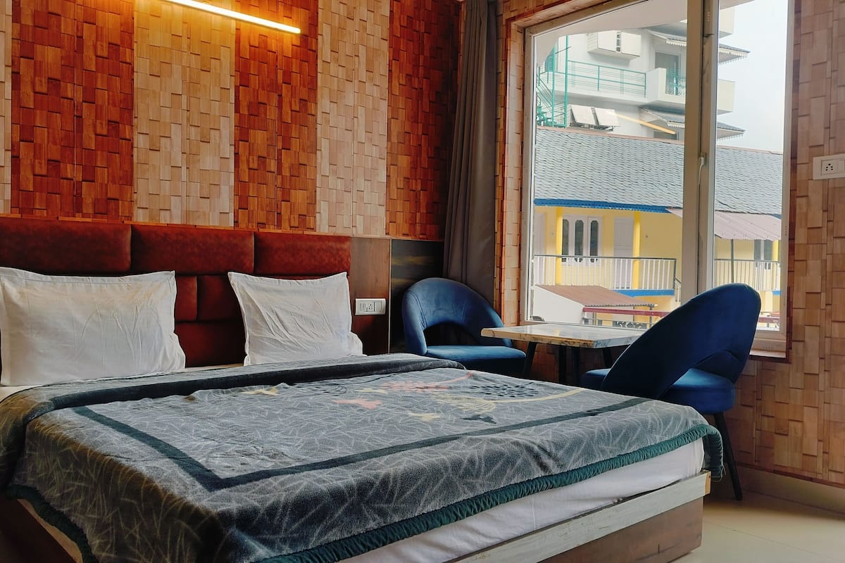Quarto de hotel com uma cama, uma mesinha, duas cadeiras e uma janela com vista para prédios.