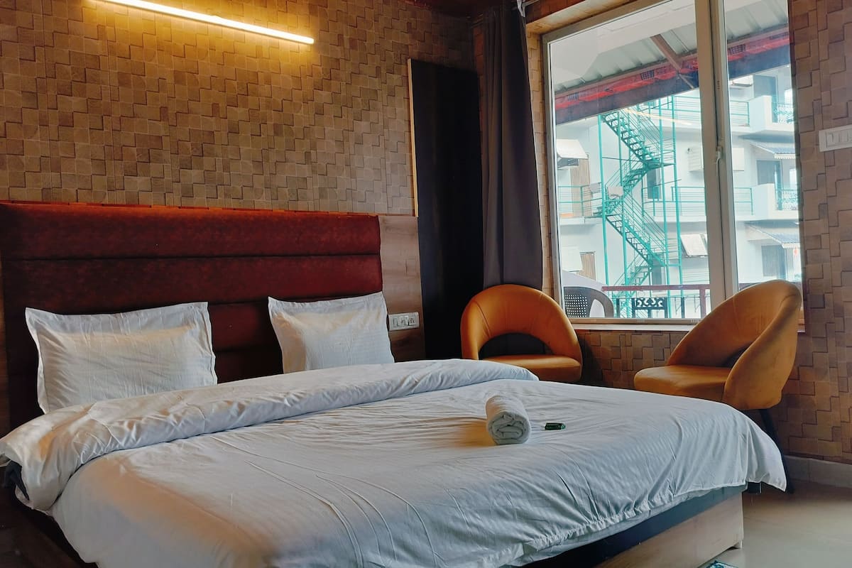 Quarto de hotel com parede em tijolos, cabeceira estofada vermelha, cama com roupa de cama branca, duas cadeiras laranja e janela com vista para um edifício.