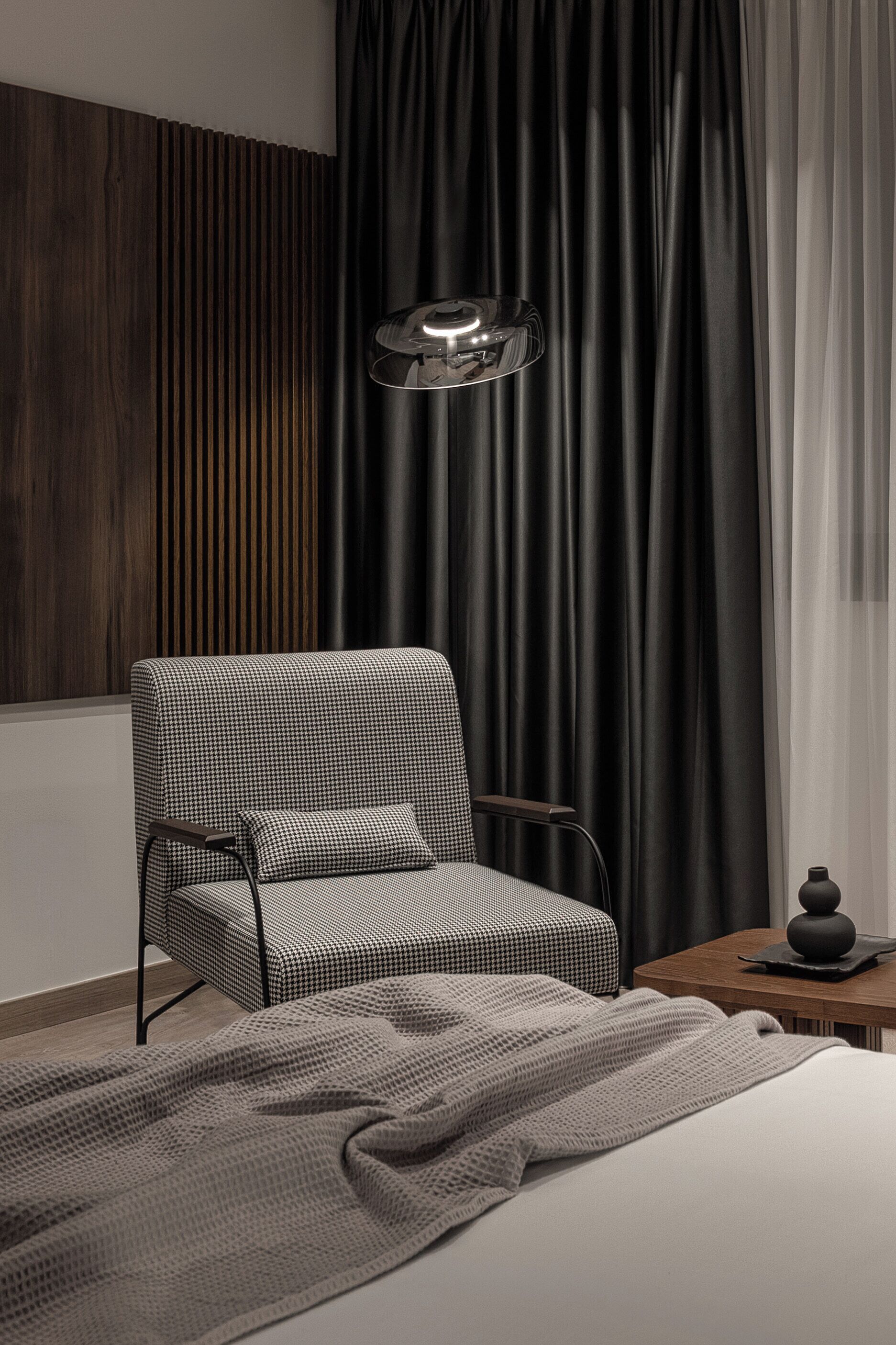 A modern bedroom with a checkered armchair, a wooden panel wall, a black curtain, and a bedside table with a lamp.