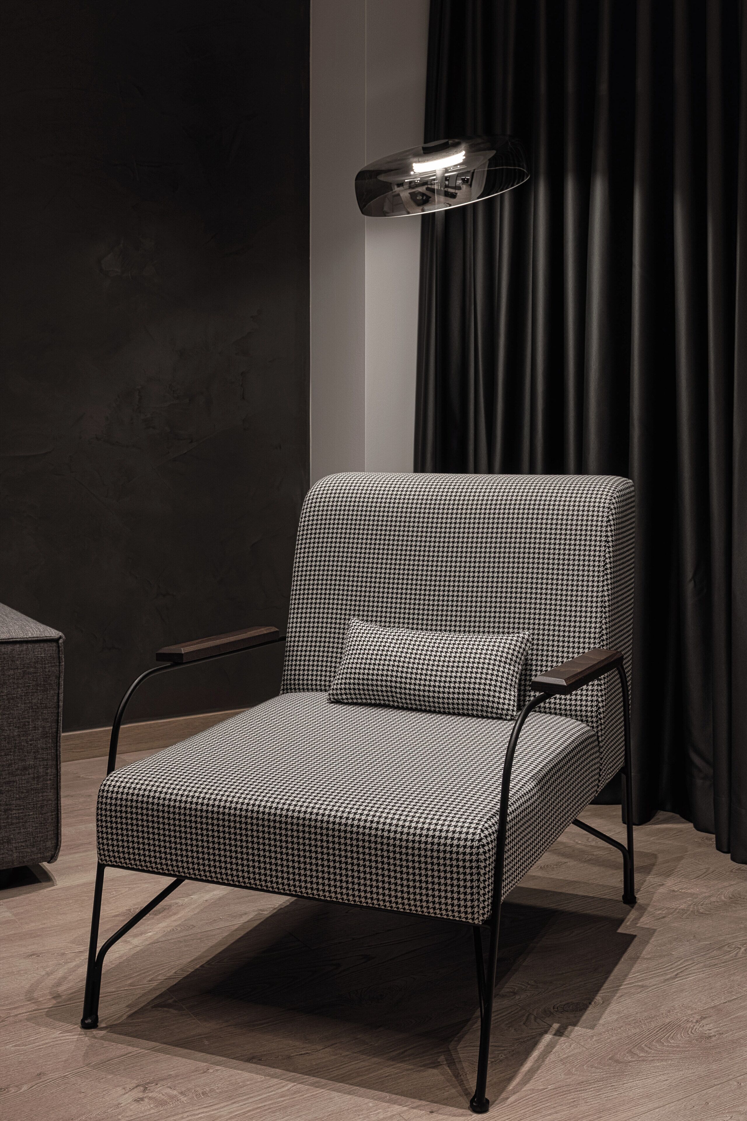 A modern chair with a checkered pattern, a side table, and a floor lamp in a room with dark curtains and a textured wall.
