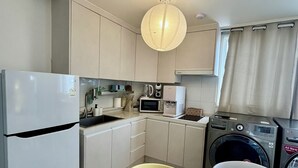 A compact kitchen with a refrigerator, microwave, toaster, and washing machine. There is a dining table with chairs and a hanging light fixture. - Gangnam Style Village (Seoul)