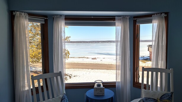 A room with a bay window overlooking a snowy landscape and a body of water. There are two white chairs with butterfly cushions, a small table with a candle and a sign, and sheer white curtains. - NEW!  The Lighthouse - Large Family-Friendly and Dog-Friendly Home on Big Platte Lake with Central A/C! (Beulah)