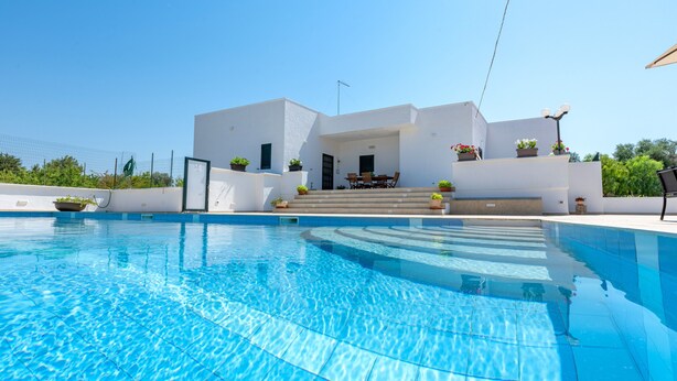 Uma área moderna à beira da piscina, com piscina azul cristalina, um edifício branco com degraus para um alpendre coberto e plantas em vasos.