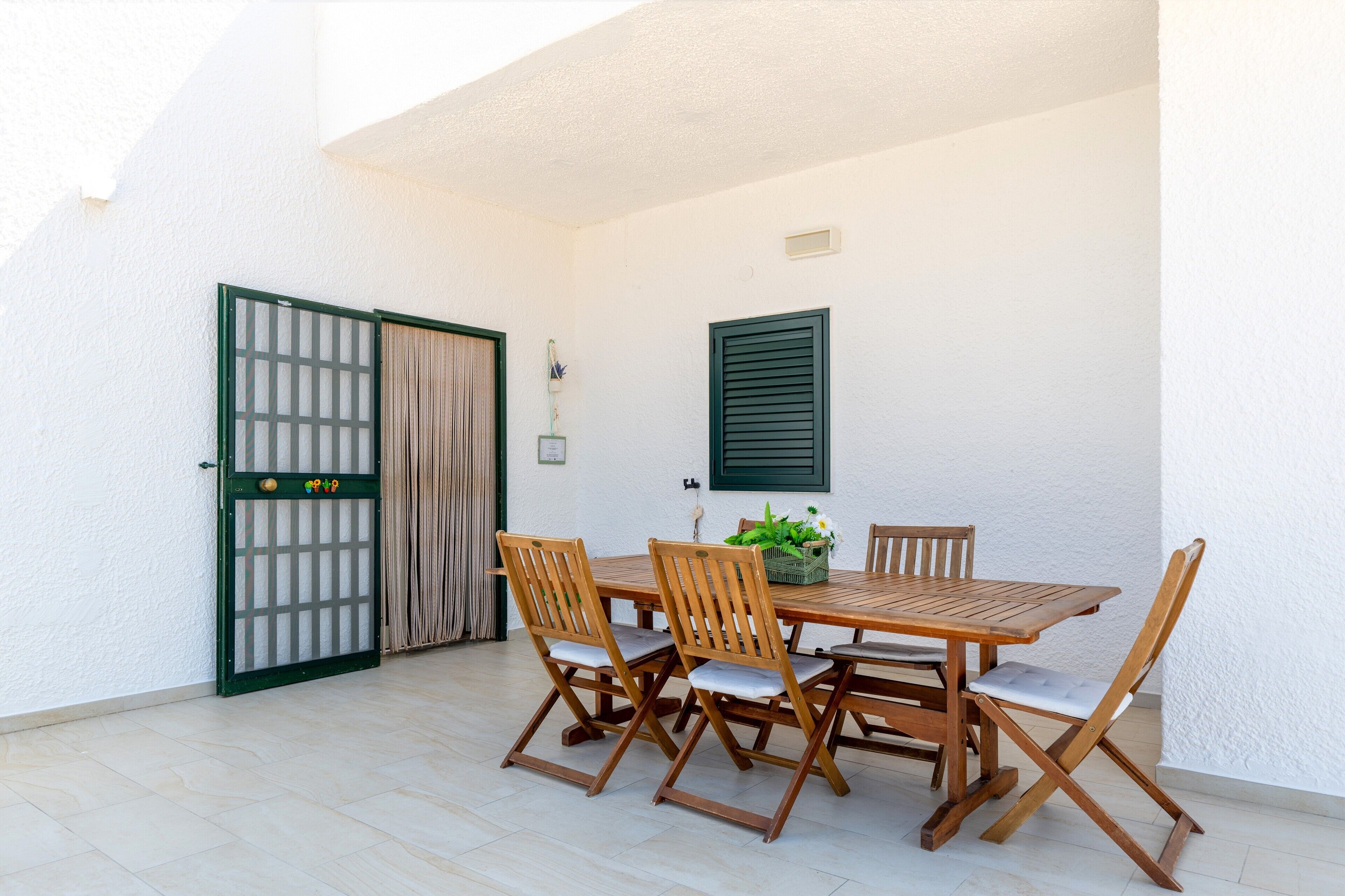 Een terras met een houten tafel en stoelen, een groen hek en een witte muur met een raam.