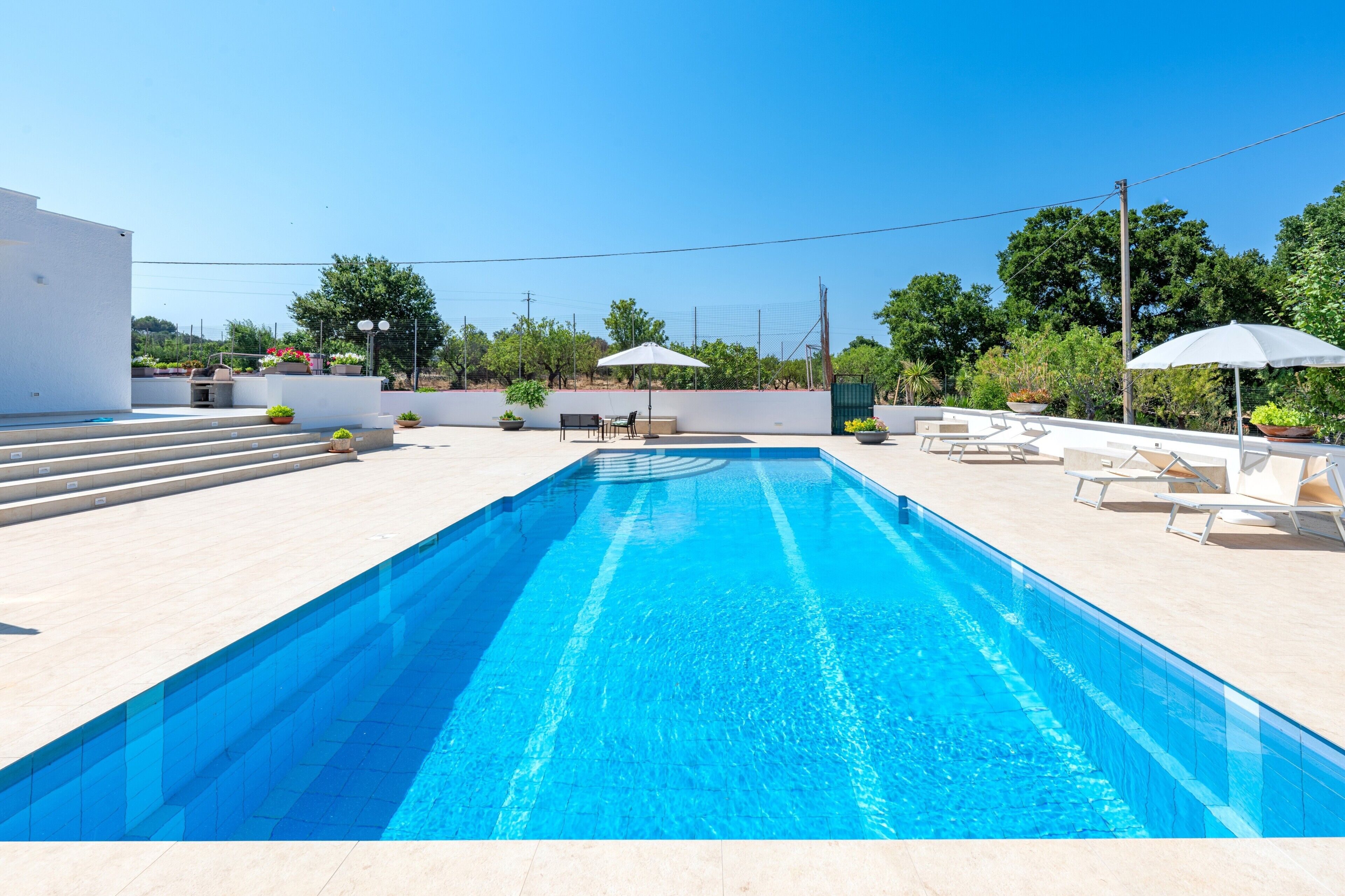 Una piscina con escalones que bajan a ella, rodeada de sillas de descanso y sombrillas.
