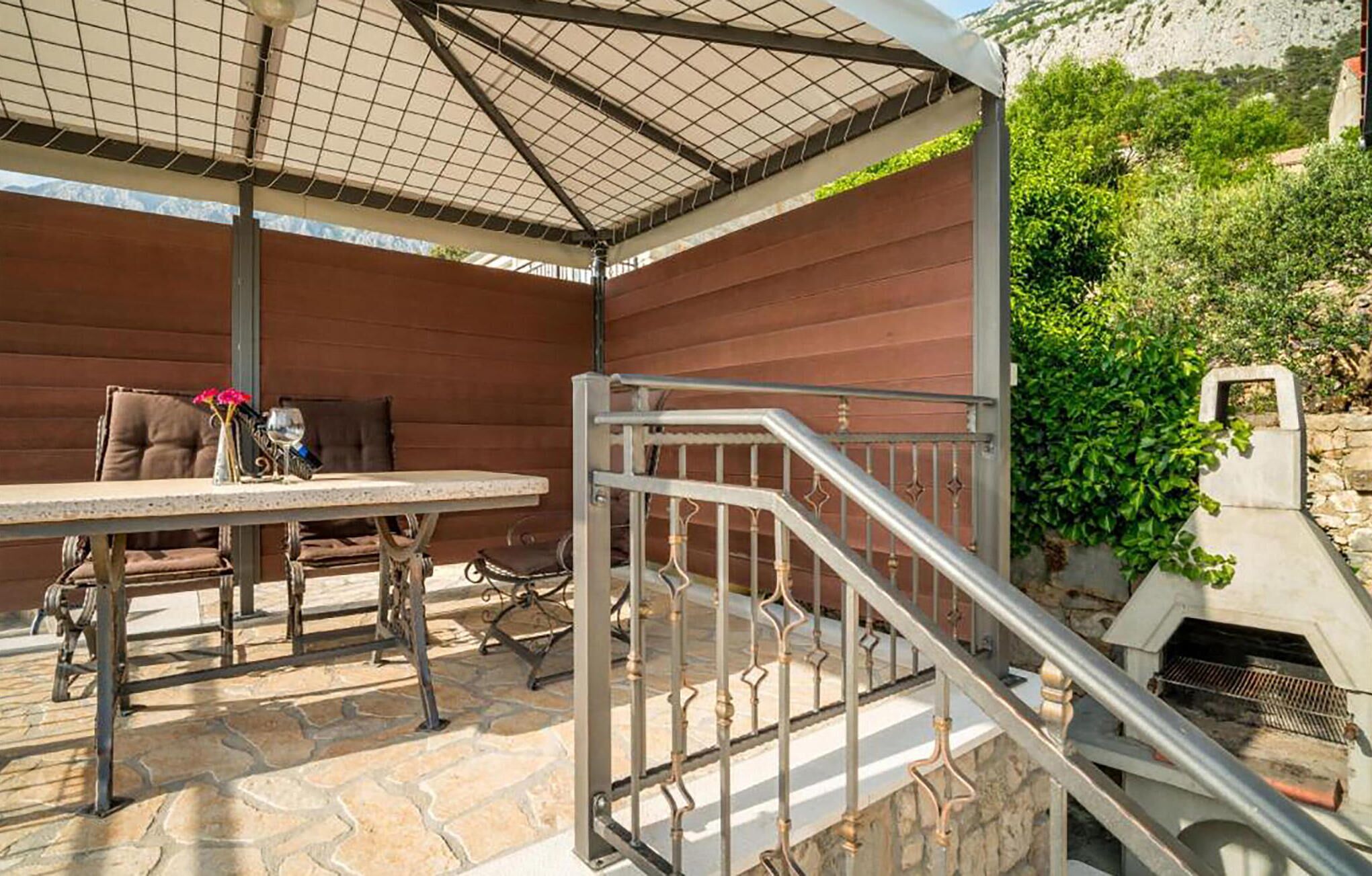 An outdoor patio with a table and chairs, a metal railing, and a stone floor.
