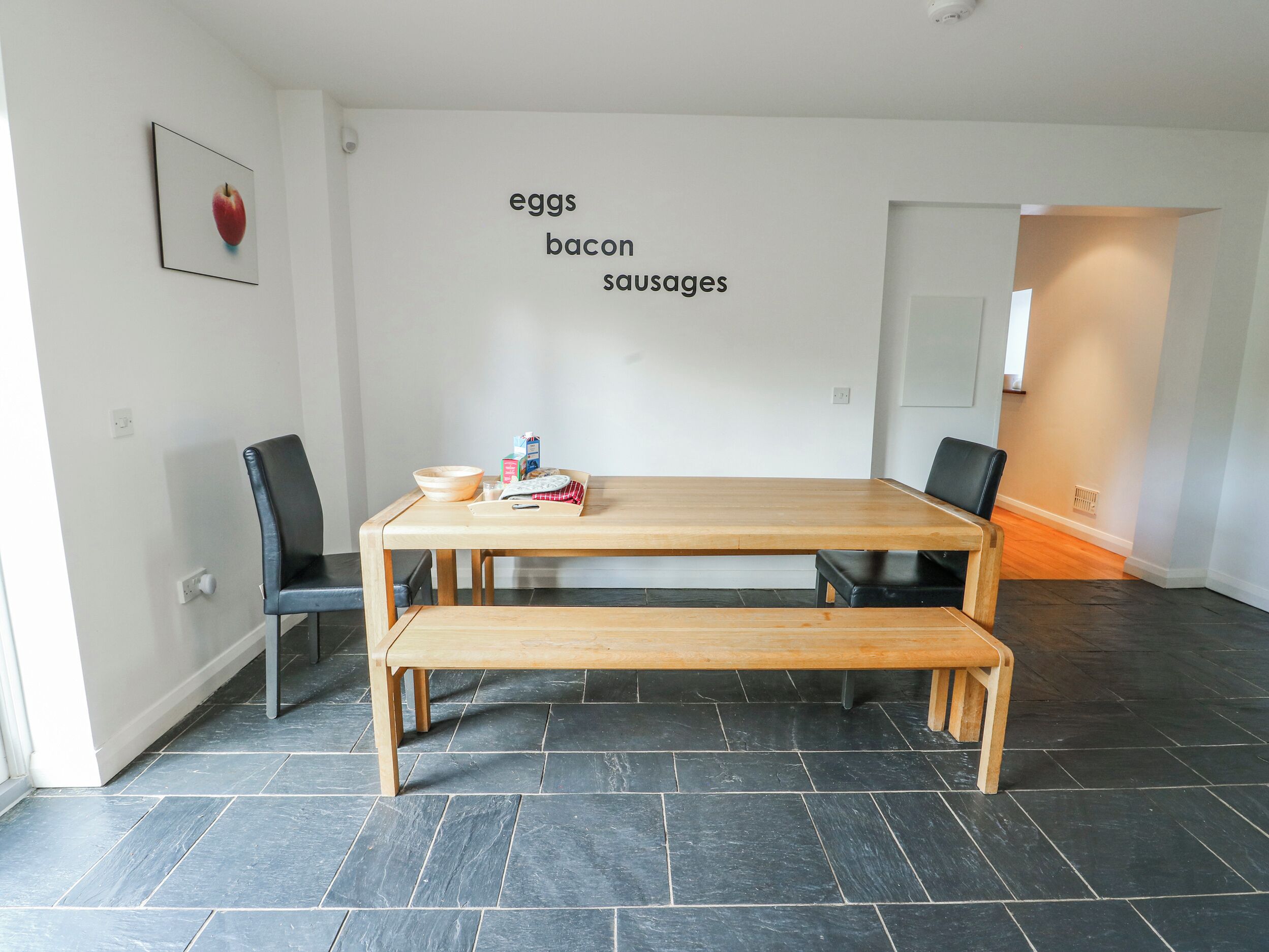 A modern dining area with a wooden table, two chairs, and a bench. A wall-mounted sign lists food items: eggs, bacon, and sausages.