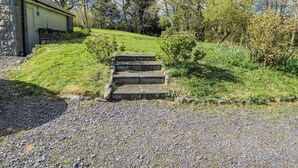 Ein Kiesweg führt zu Steinstufen, einer grasbewachsenen Hügelkuppe und einem Gebäude mit einer Stein-Fassade.