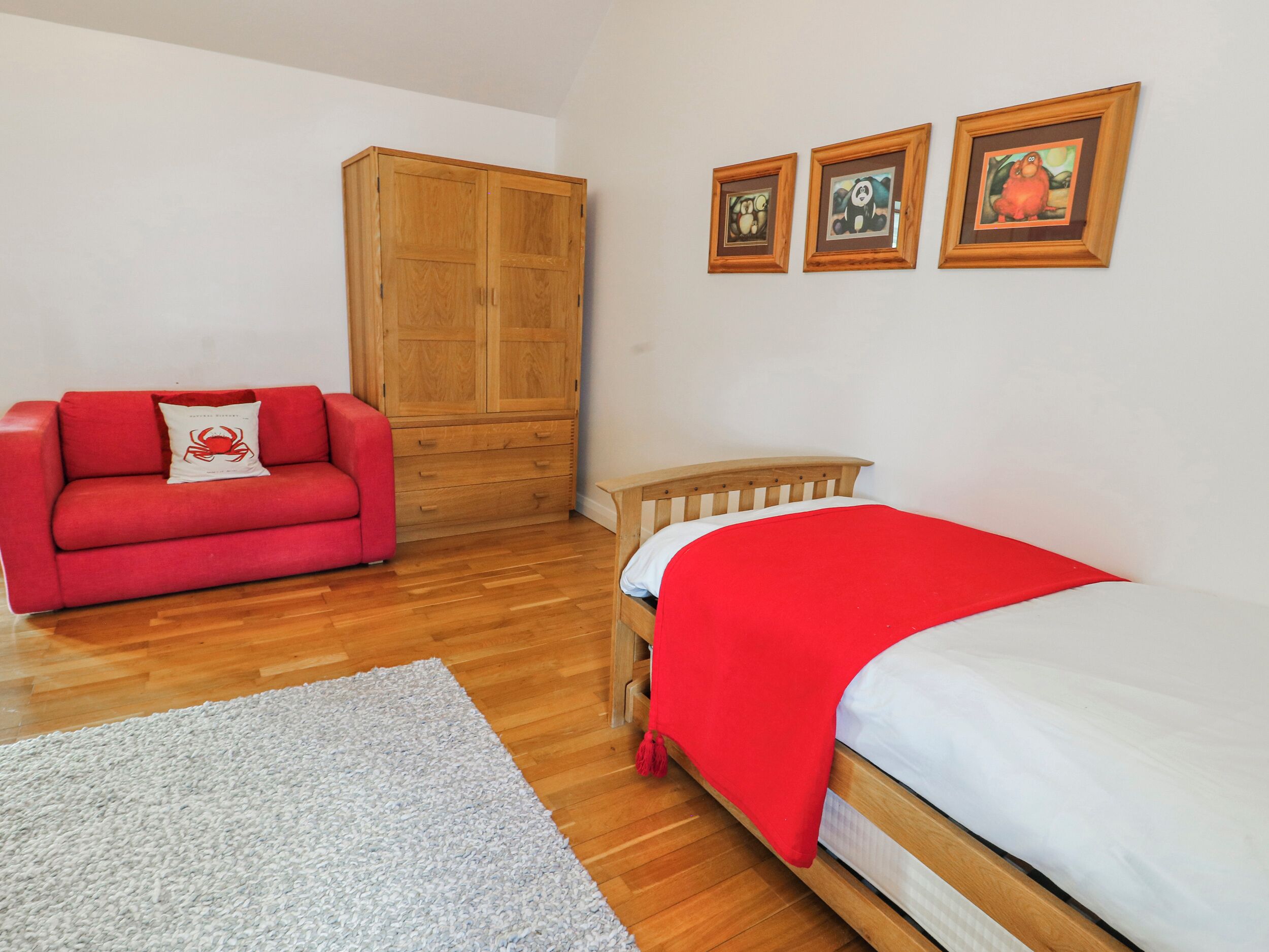 Un dormitorio con un armario de madera, un sofá rojo, una cama con manta roja, una alfombra gris y tres cuadros enmarcados en la pared.