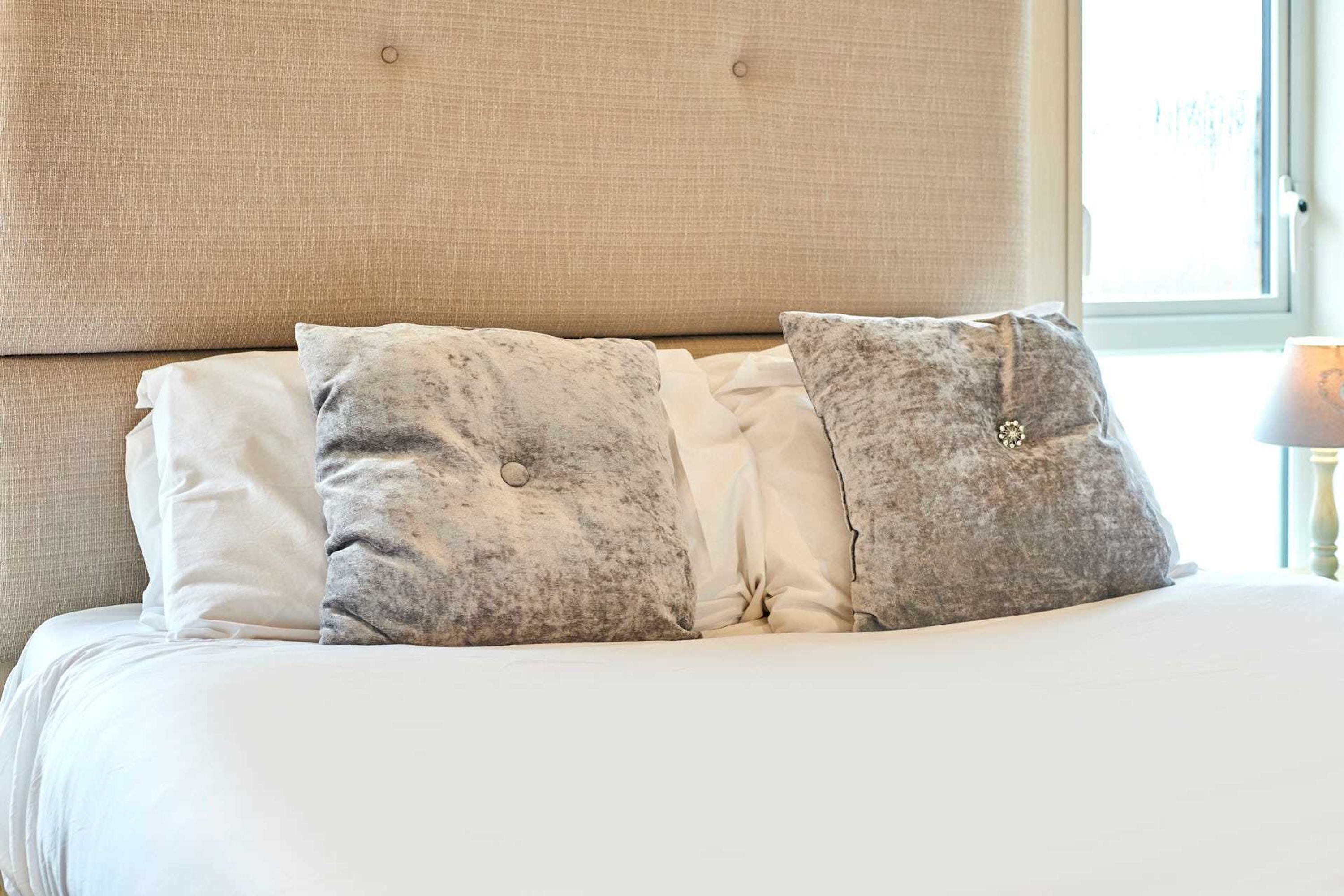 A bed with a beige upholstered headboard, white and grey pillows, and a lamp on a bedside table.