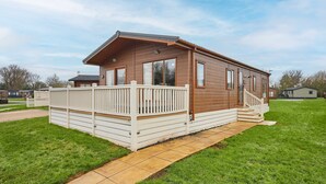 A single-story mobile home with a wooden exterior, a white porch with a railing, and a paved driveway. - Host Stay Rowan Lodge (Malton)
