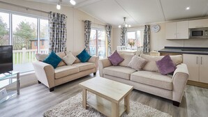 A spacious living room with two sofas, a coffee table, and a TV on a glass stand. A kitchen area is visible with cabinets and a microwave. - Host Stay Rowan Lodge (Malton)