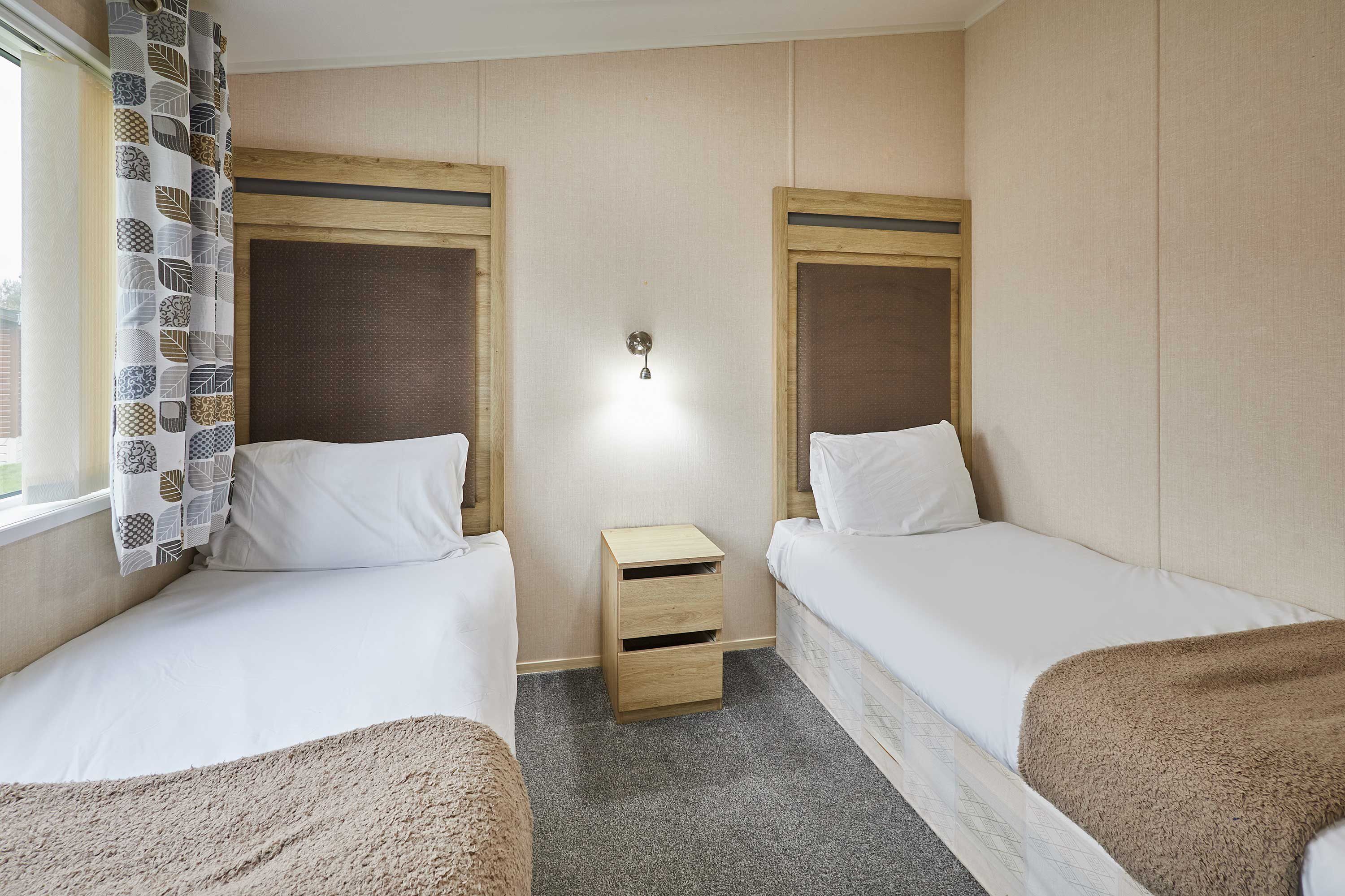 A hotel room with two single beds, a small wooden nightstand, and a wall-mounted lamp.