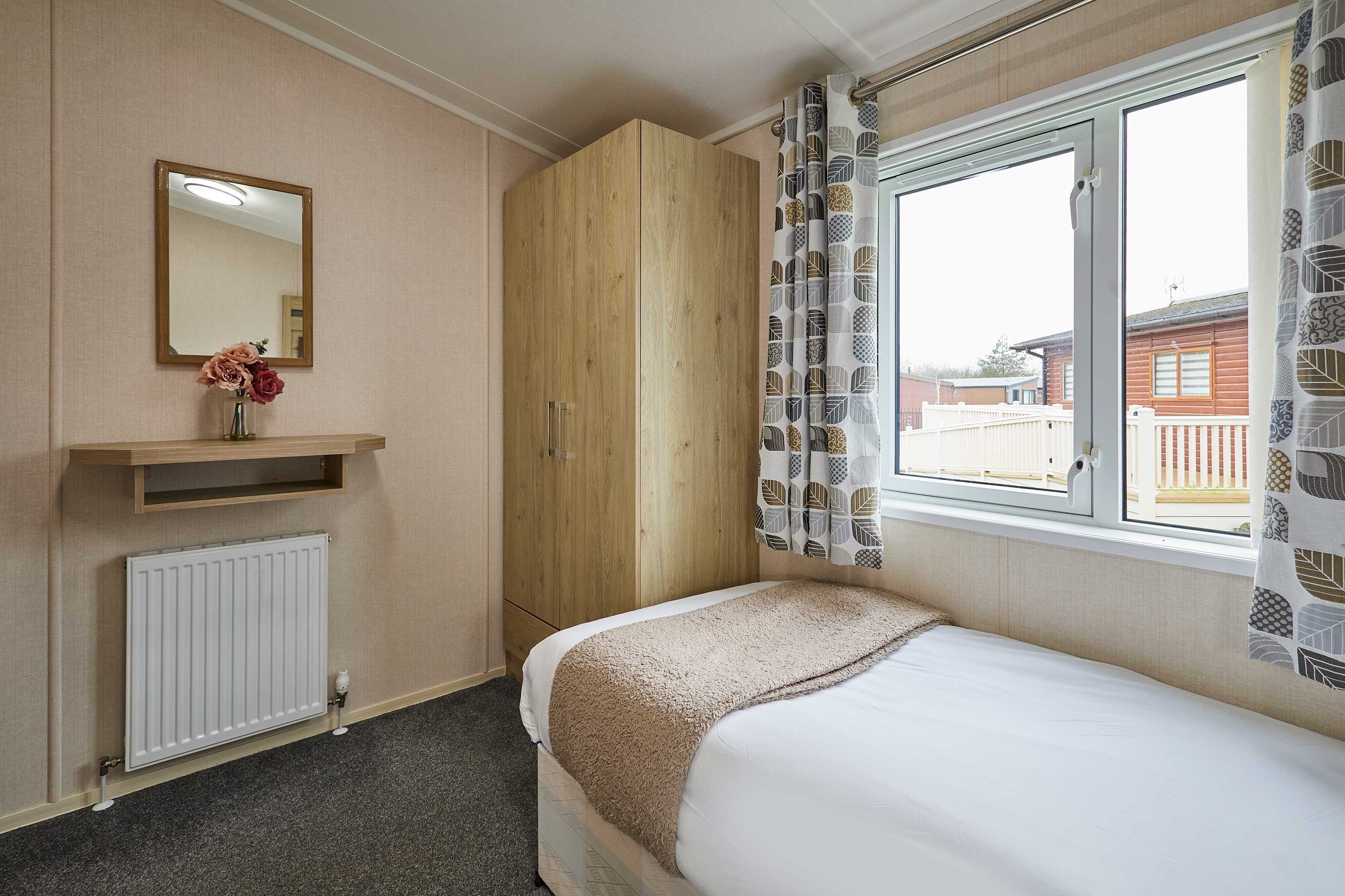 A bedroom with a bed, a wooden wardrobe, a radiator, a mirror, and a window with patterned curtains.