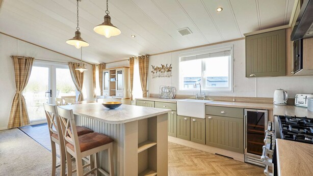 A kitchen with a central island, bar stools, a stove, and a window overlooking a deck. - Host Stay Hedgerow Lodge (Malton)