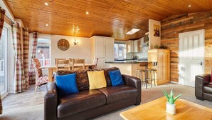 A cozy living room with a leather sofa, wooden coffee table, and a dining area with a table and chairs. A kitchen with white cabinets and a bar area is visible in the background. - Host Stay Crystal Lodge (Malton)