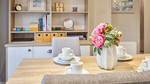 Ein Essbereich mit einem Holztisch, der für Tee gedeckt ist, einer Vase mit rosa Blumen und einem Bücherregal im Hintergrund.
