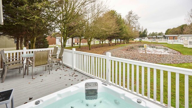 Ein Whirlpool auf einer Holzterrasse mit weißer Geländer, umgeben von Bäumen und einer Rasenfläche.