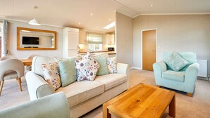 A living room with a sofa, a coffee table, and a light blue armchair. There is a kitchen in the background with cabinets and a microwave. - Host Stay Deneb Lodge (Malton)