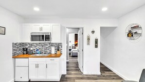 Une cuisine moderne avec des armoires blanches, un four à micro-ondes et un plan de travail en bois. On y trouve un coin repas avec une table et des chaises, ainsi qu'un couloir menant à une chambre.