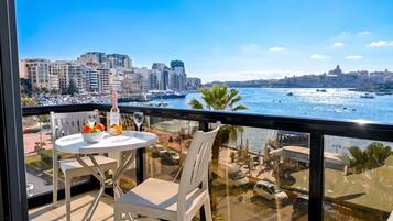 A balcony with a table set for two, overlooking a marina with boats and a cityscape.