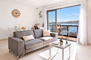 A modern living room with a grey sofa, a coffee table, and a view of the sea through large windows. - A Sunny Seaview In Central Sliema (Sliema)