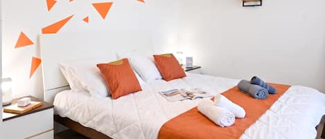 Una cama bien hecha con una colcha blanca y naranja, almohadas y una revista. Hay dos mesitas de noche con lámparas y una planta pequeña en una de ellas.