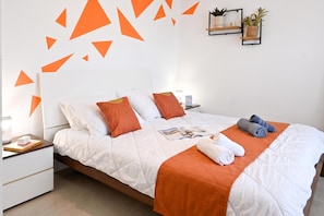 A neatly made bed with a white and orange bedspread, pillows, and a magazine. There are two bedside tables with lamps and a small plant on one. - A Sunny Seaview In Central Sliema (Sliema)