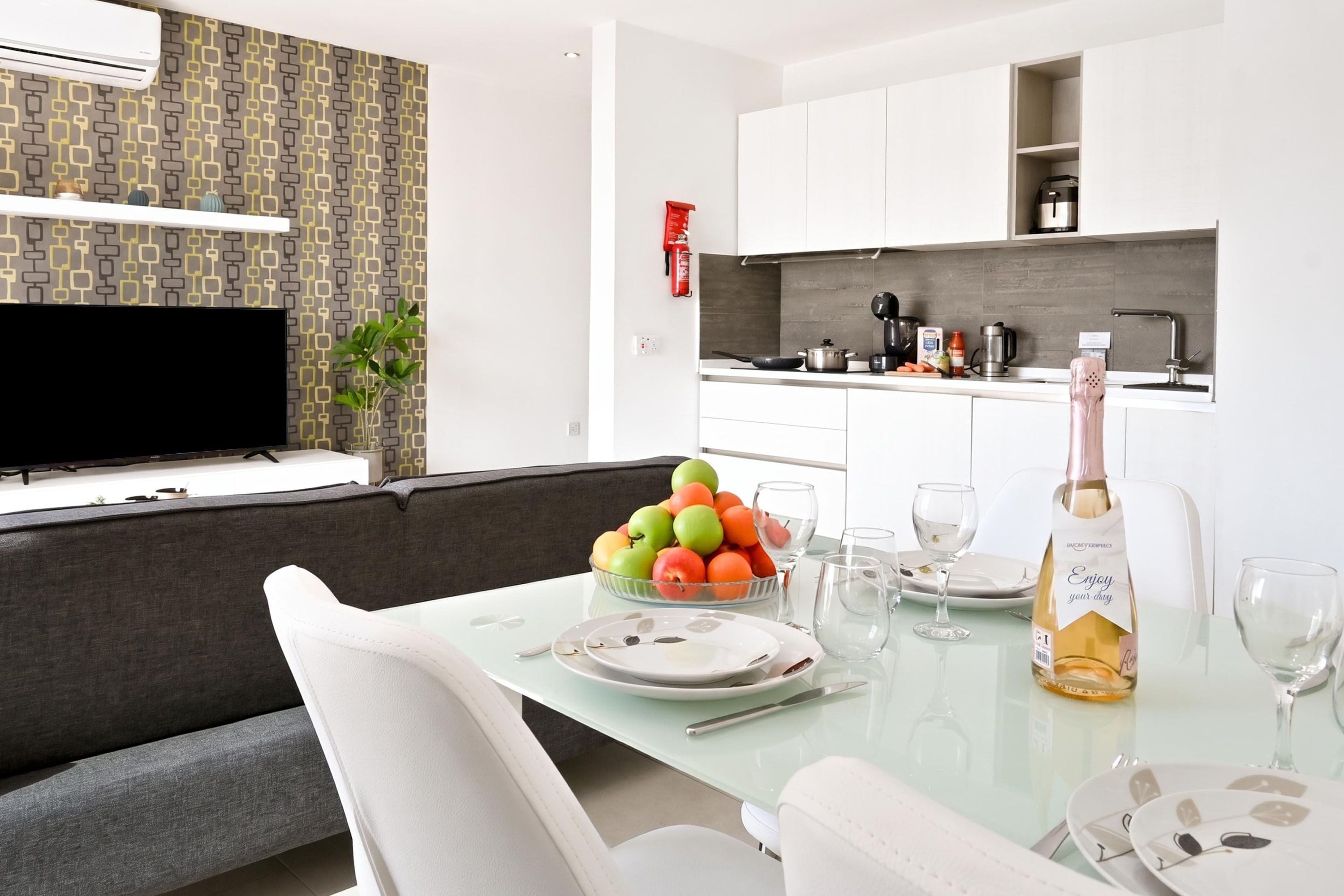 A modern living room with a sofa, a glass dining table set for two, and a TV mounted on the wall.