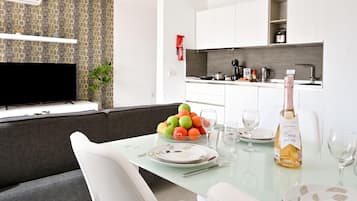 A modern living room with a sofa, a glass dining table set for two, and a TV mounted on the wall.