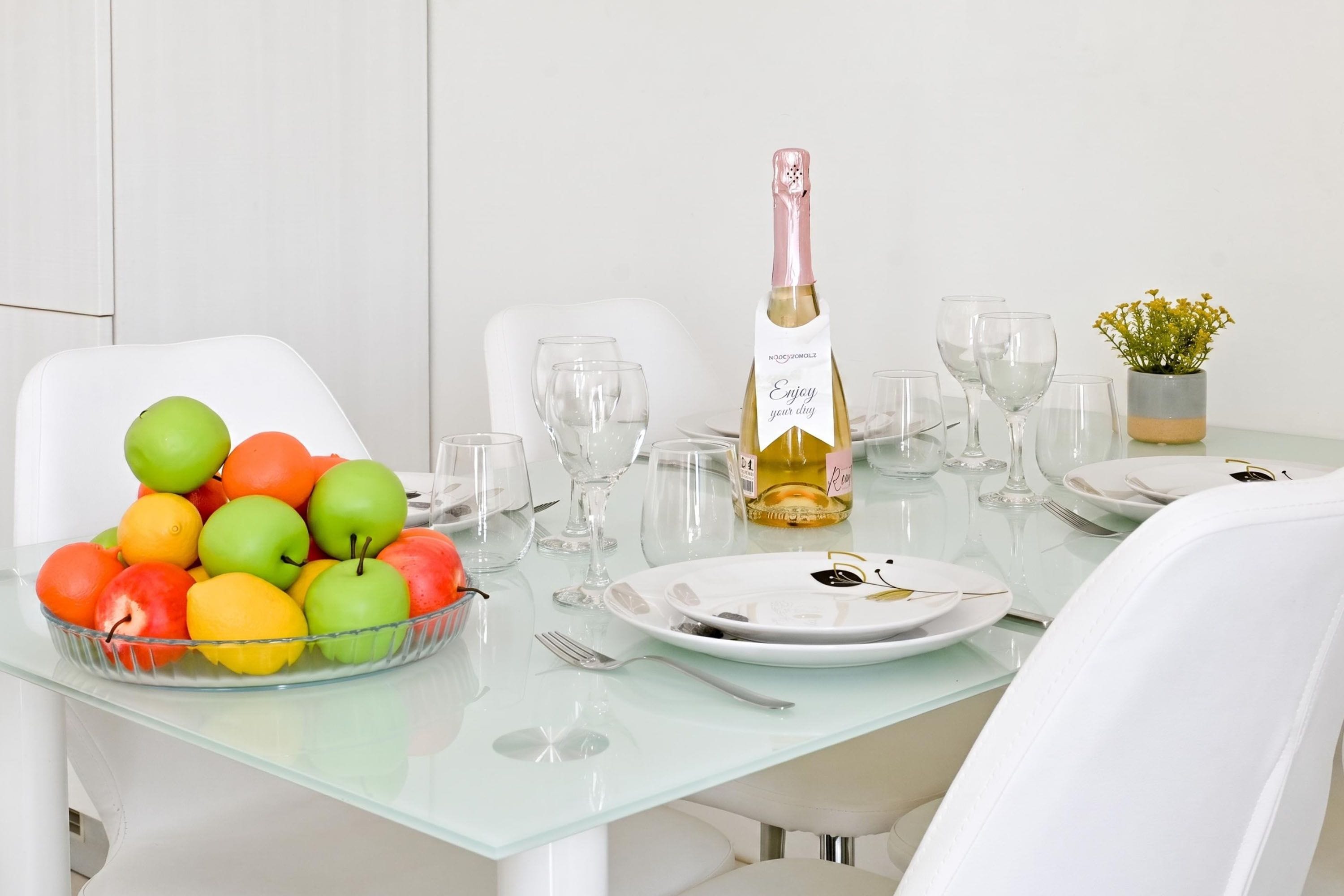 A dining table set with a bottle of white wine, clear glasses, a bowl of fruit, and a small potted plant.