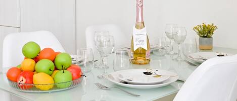 A dining table set with a bottle of white wine, clear glasses, a bowl of fruit, and a small potted plant.