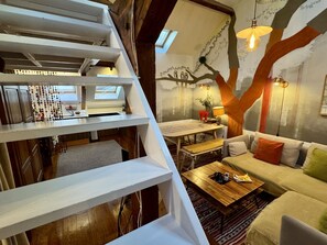 Ein mehrgeschossiges Interieur mit Treppe, Essbereich, einer Couch und Wandgemälde.