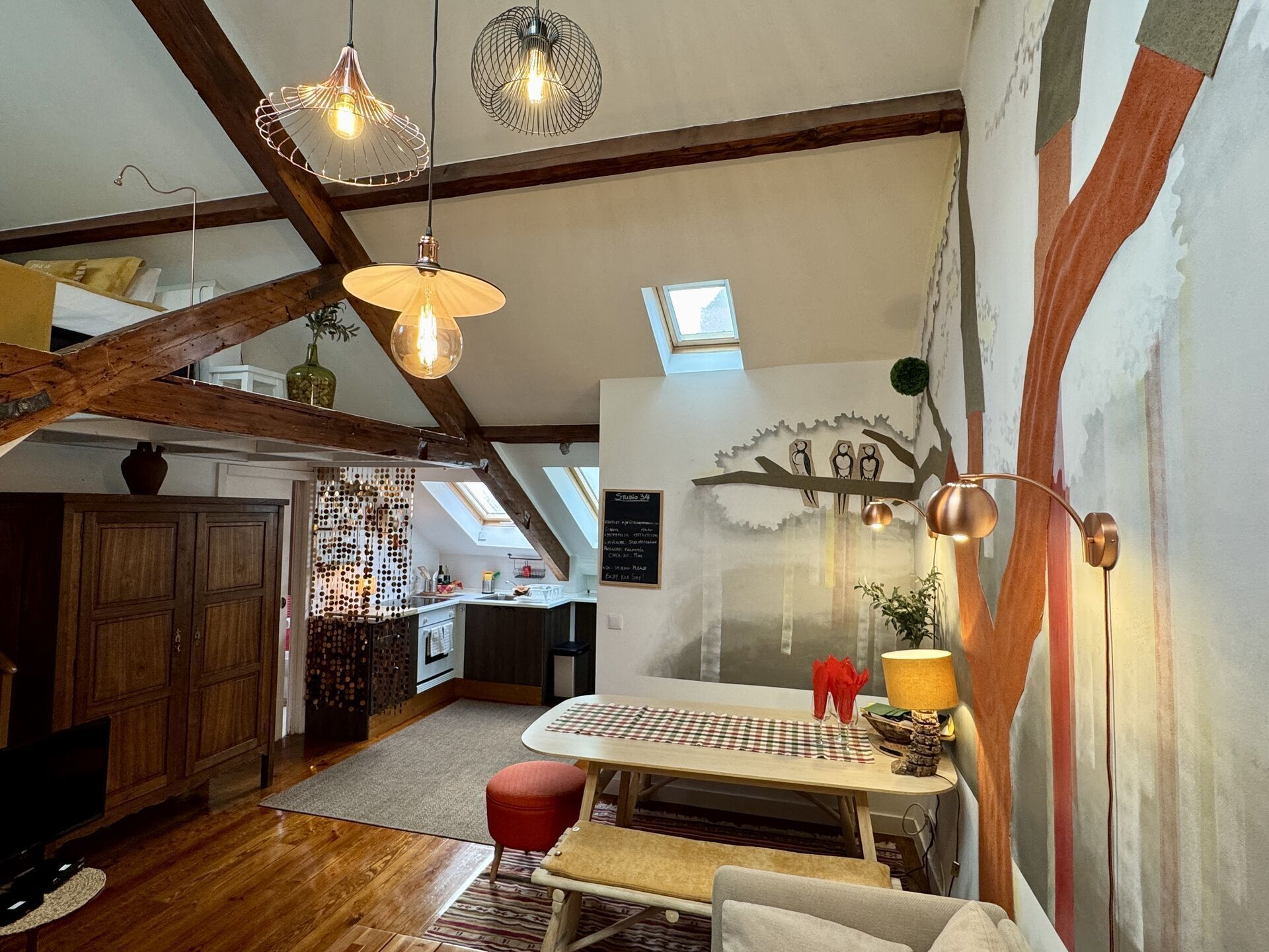 A loft-style room with exposed wooden beams, a dining area with a checkered tablecloth, and a mural on the wall.