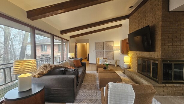 A spacious living room with a fireplace, large windows, and a television mounted on the wall. - Serene 4-bedroom house with brook and forest in charming Racine (Racine)