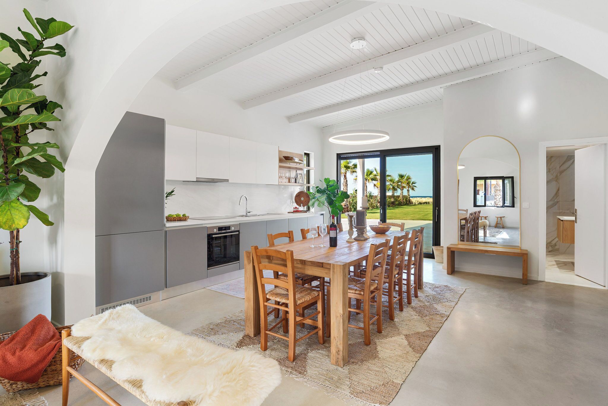 Una cocina moderna con armarios blancos, un comedor con mesa y sillas de madera, y un gran ventanal con vistas a palmeras.