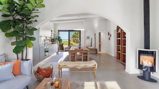 Een moderne woonkamer met een bank, een lange hocker en een open haard. Er is een eethoek met een tafel en stoelen, en een keuken op de achtergrond.