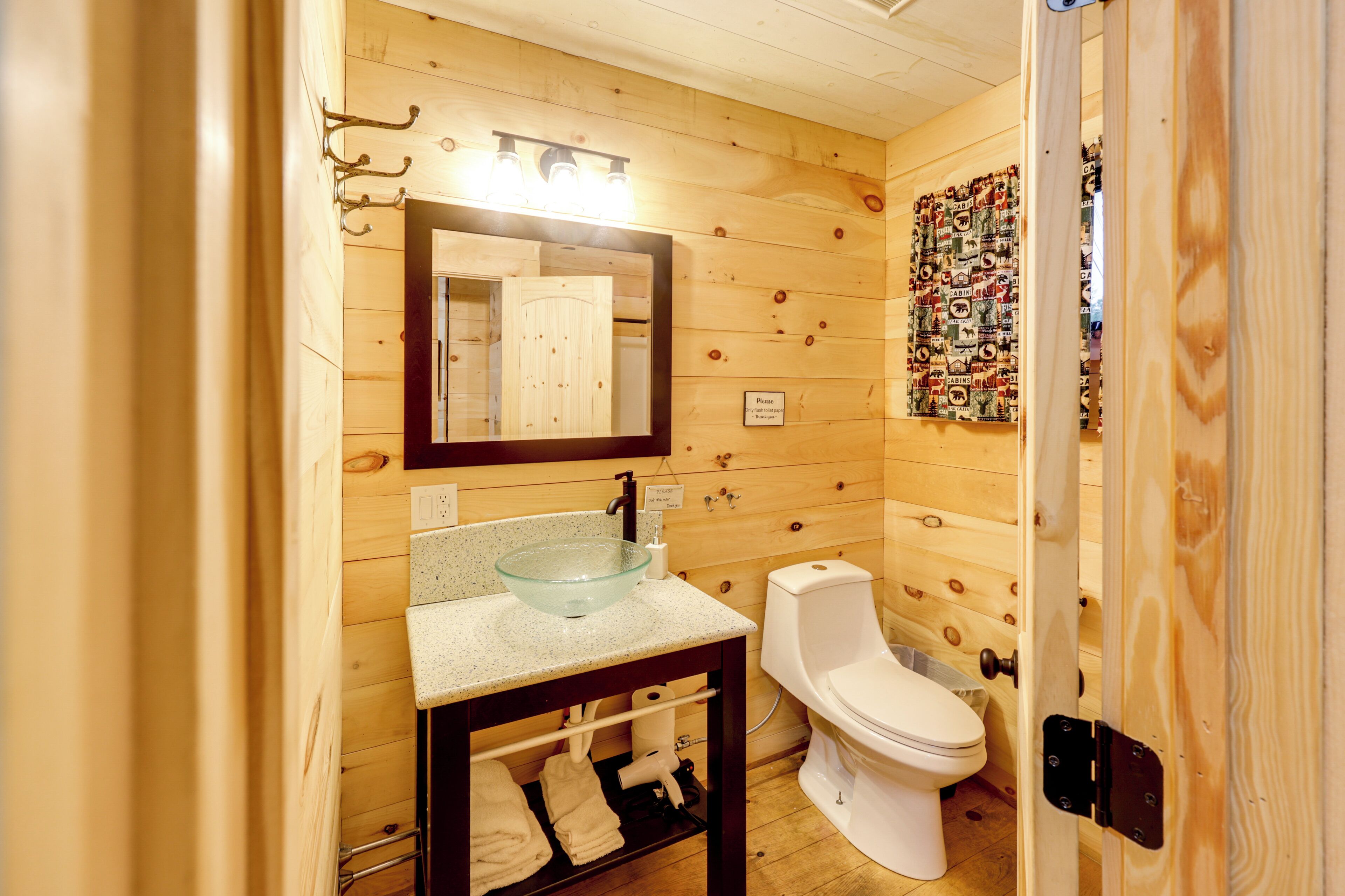 Une salle de bain aux murs lambrissés, dotée d’un lavabo en verre, de toilettes et d’un miroir placé au-dessus du lavabo.
