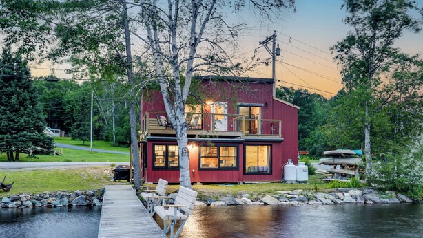 Une cabane rouge avec un quai s’avançant dans un lac paisible, entourée d’arbres et sous un ciel dégagé.