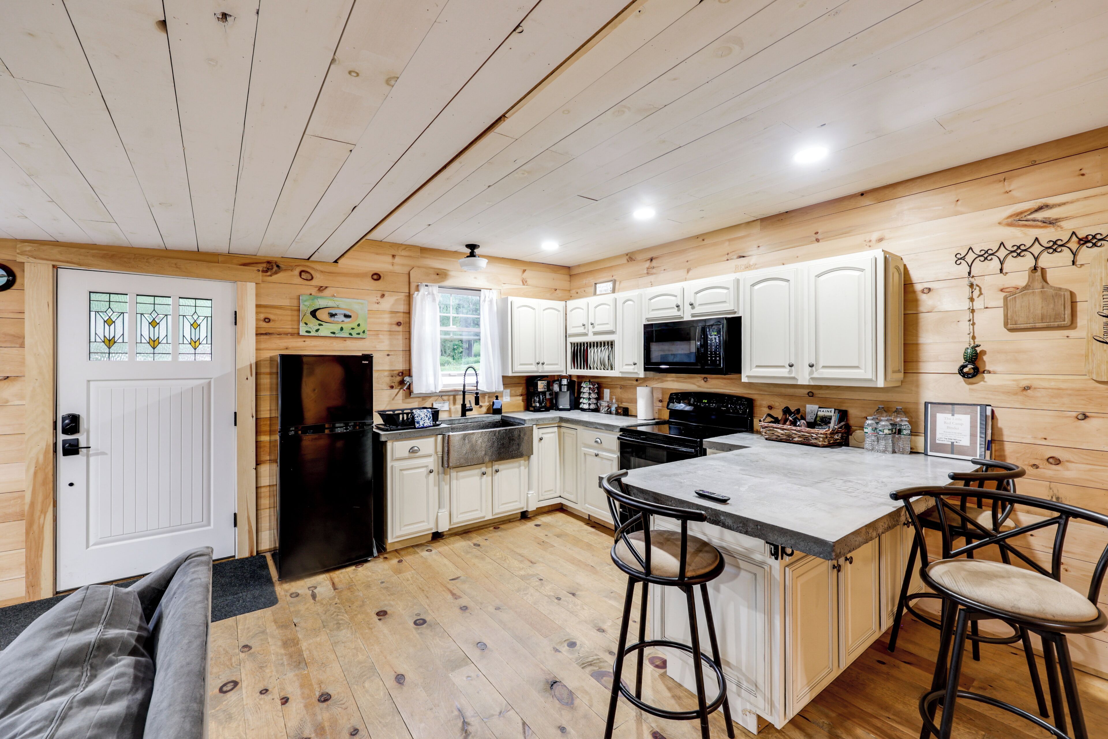 A kitchen with wooden walls, white cabinets, a black refrigerator, a microwave, a sink, and a bar area with stools.