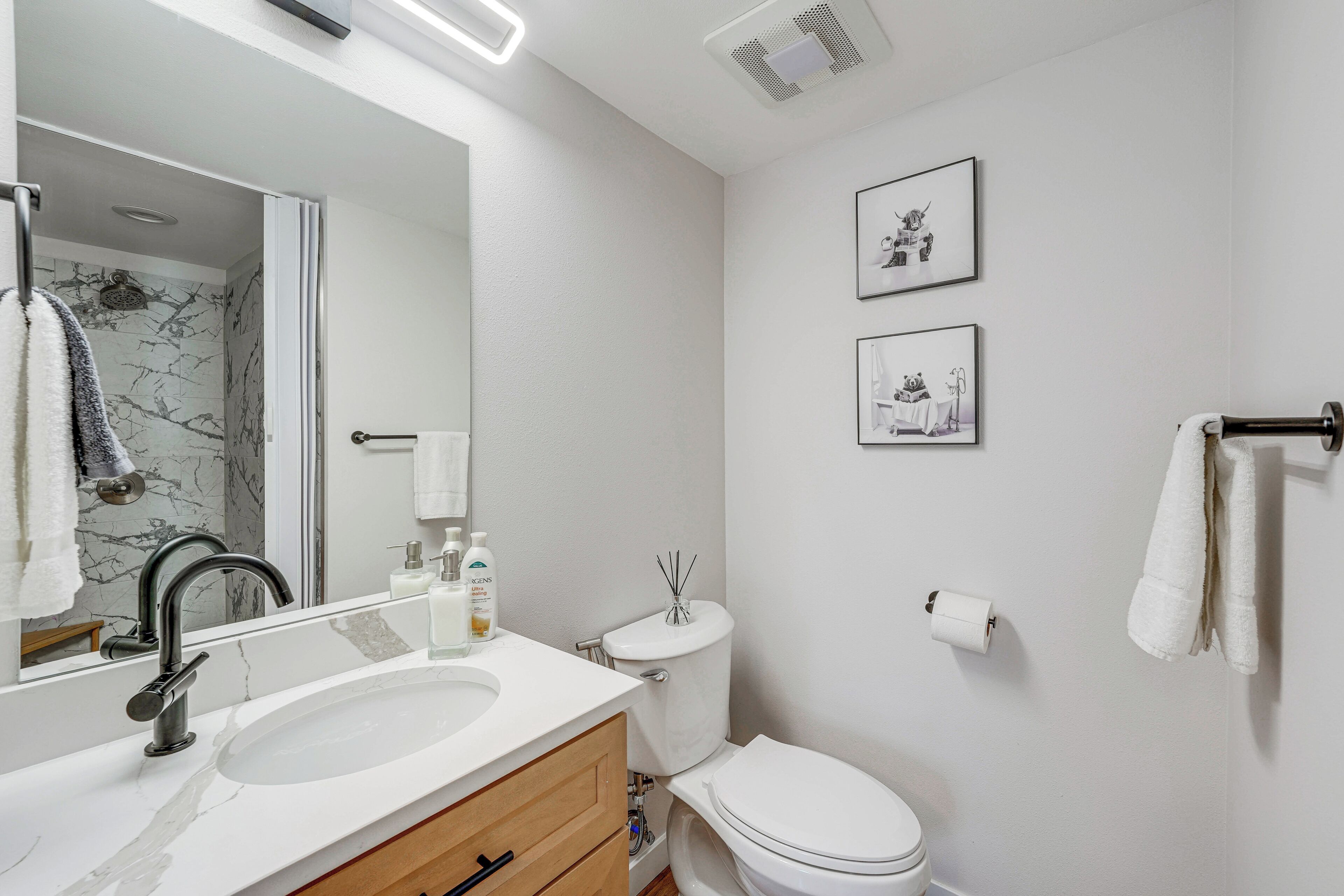 A modern bathroom with a white sink, mirror, and toilet. There are two framed pictures on the wall, a towel rack, and a toilet paper holder.