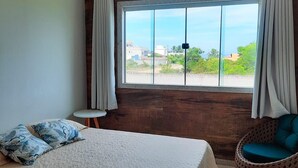 A bedroom with a large bed, a small side table, a wicker chair, and a window with curtains. - Casa Sol e Sal is the beach house with a pool for your family. (Prado)