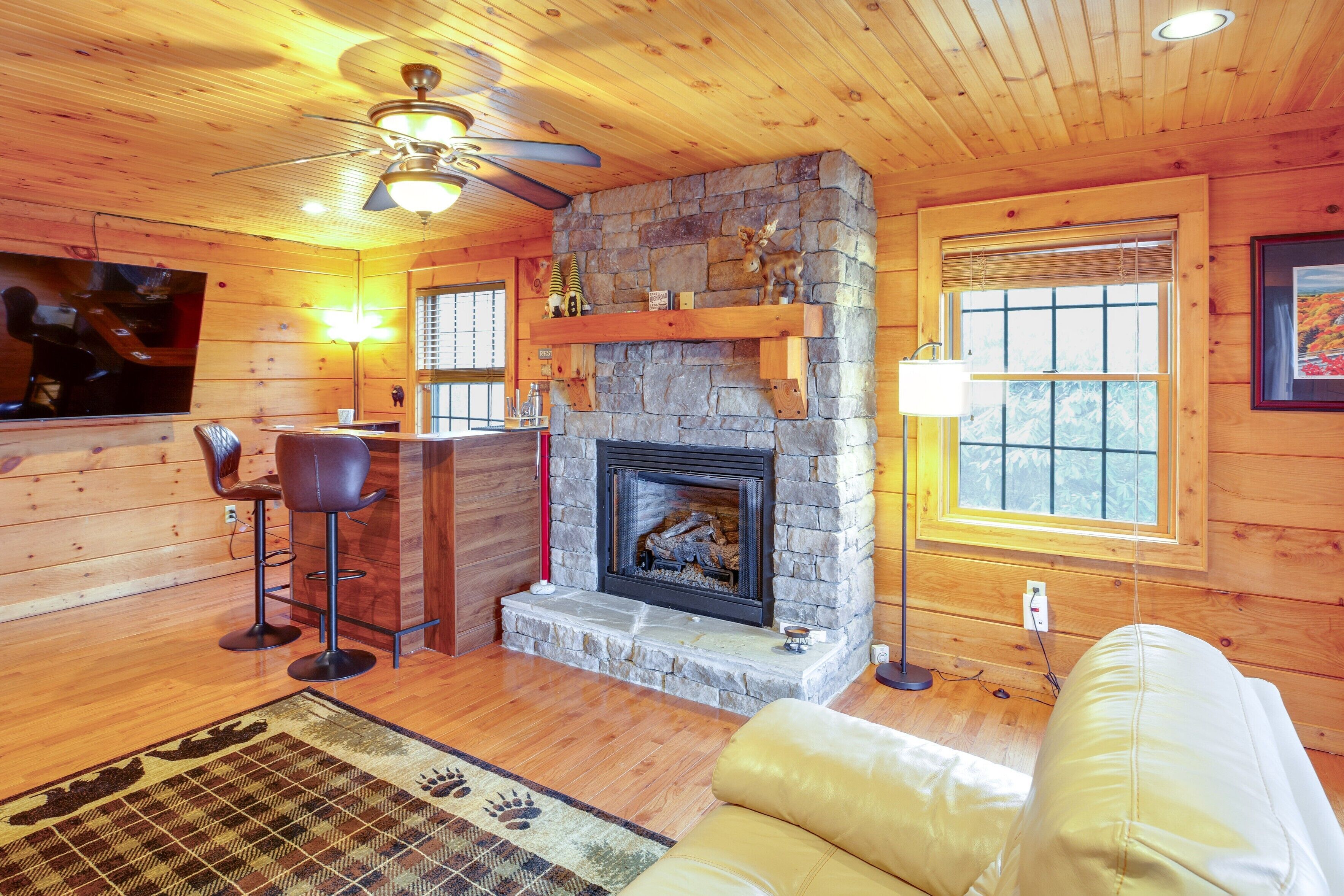 Un intérieur de chalet chaleureux avec une cheminée en pierre, des murs en bois, un ventilateur de plafond, un coin bar et un canapé en cuir.