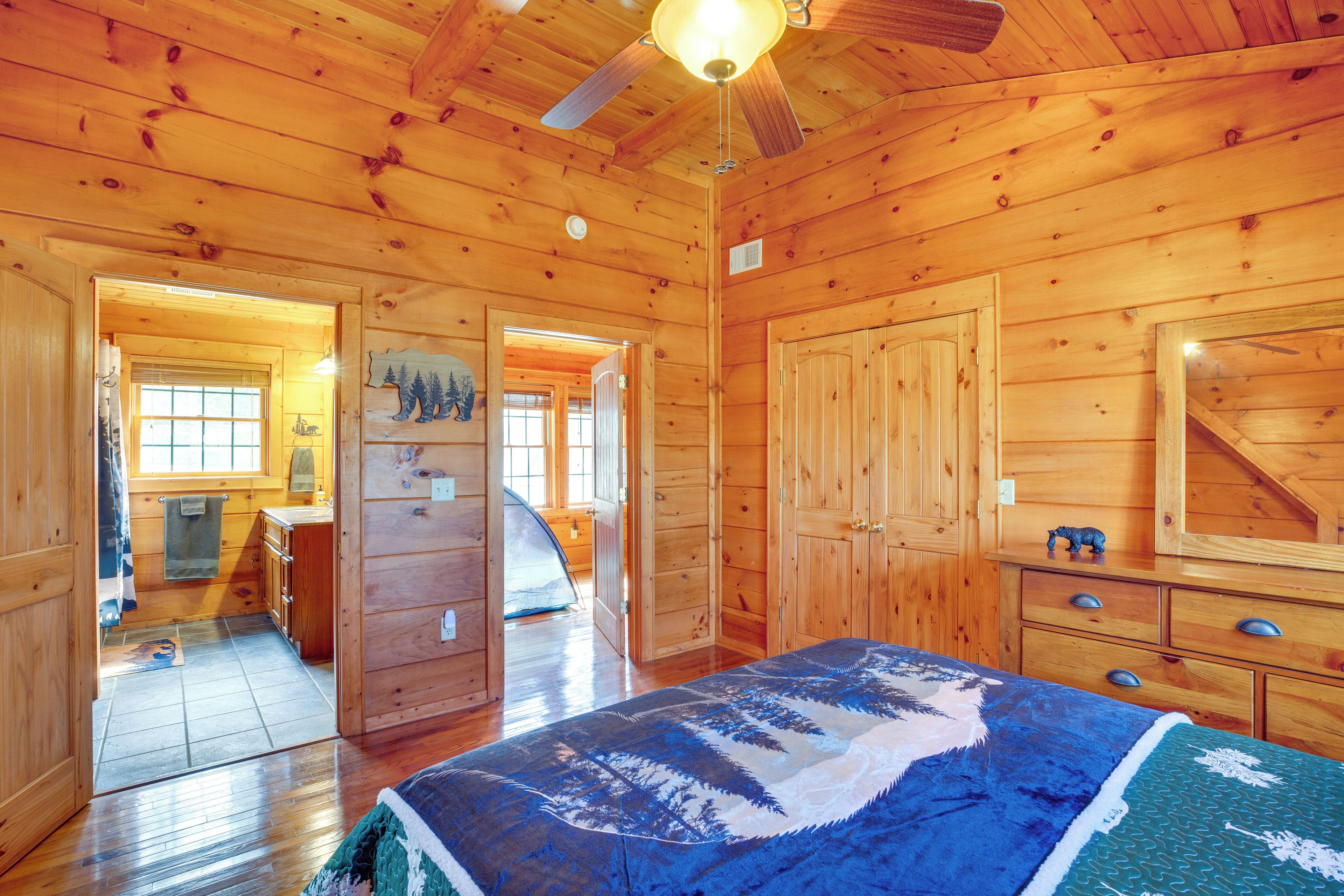 Interior de una cabaña de madera con una cama, armarios de madera, un ventilador de techo y una puerta que da a otra habitación.