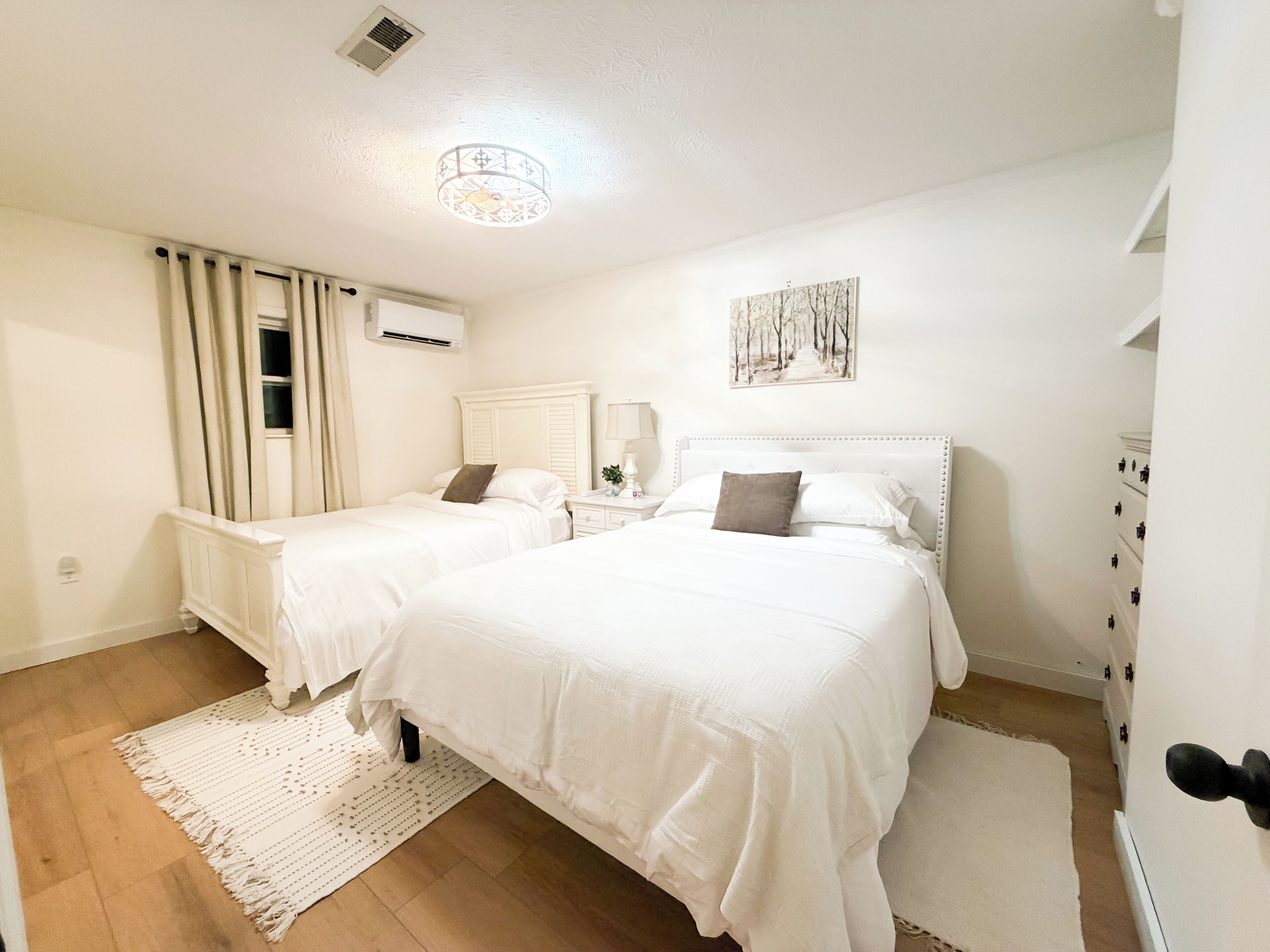 A bedroom with two beds, a bedside table with a lamp, a wall-mounted air conditioner, and a painting.