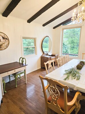 A dining area with a white table, wooden chairs, a chandelier, and a large window with a view of greenery.
