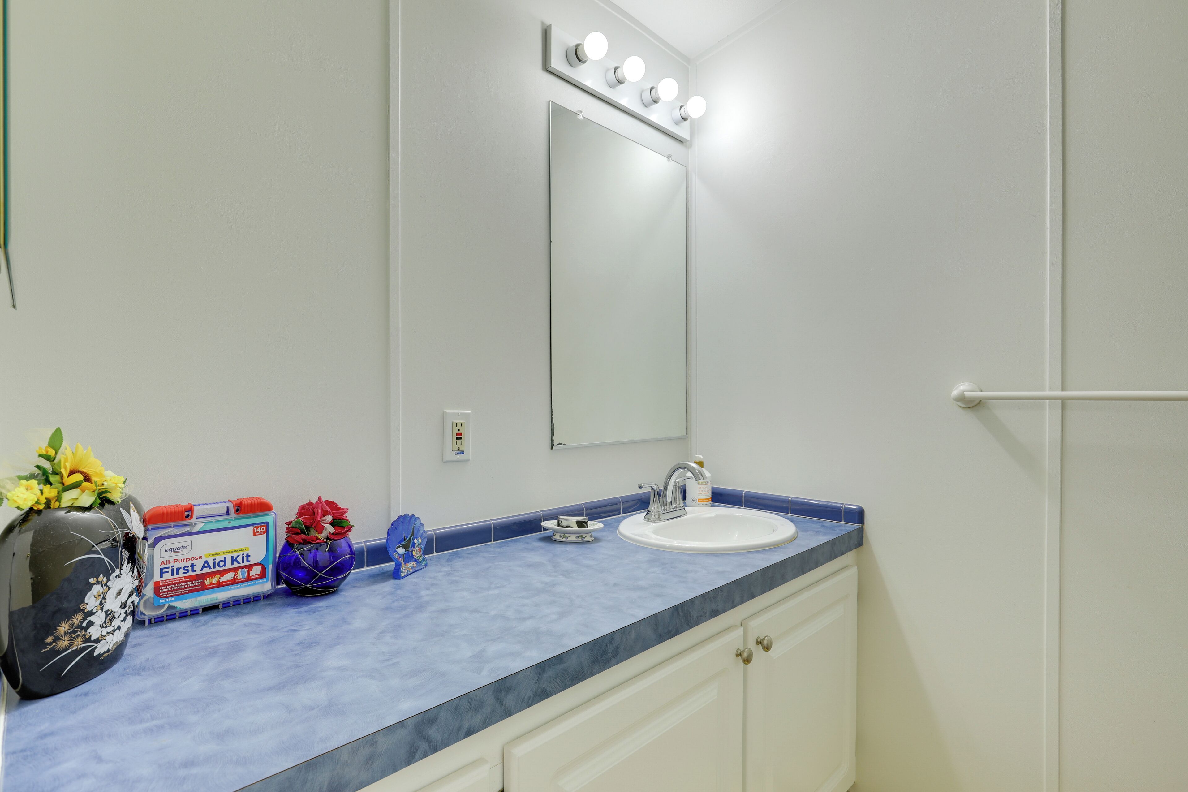 A bathroom with a white sink, blue countertop, and a mirror with lights. There is a first aid kit and a vase with flowers on the counter.