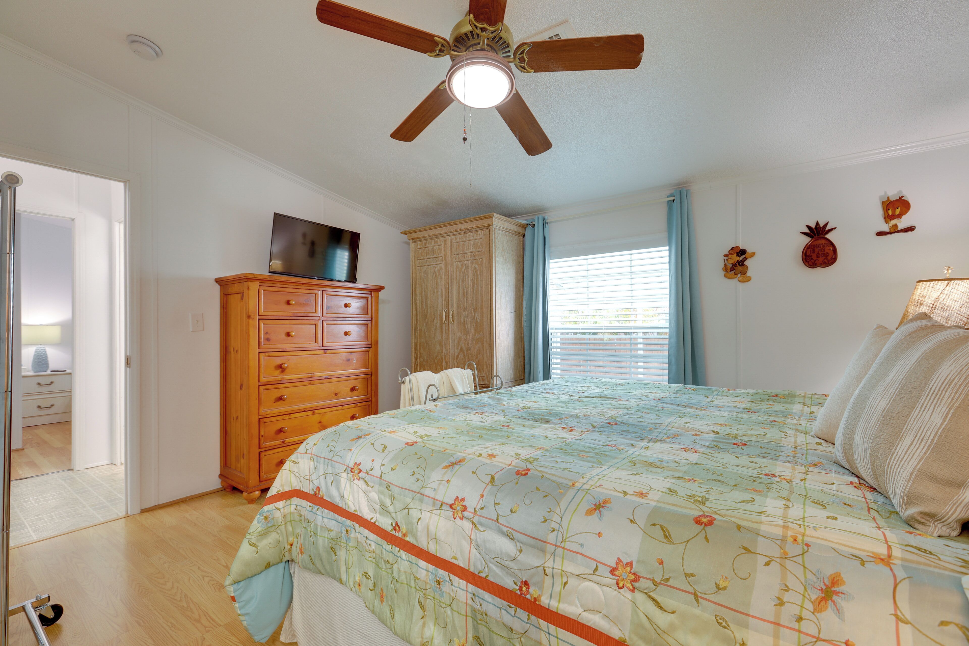 A bedroom with a wooden dresser, a bed with floral bedding, a ceiling fan, and a window with blinds.